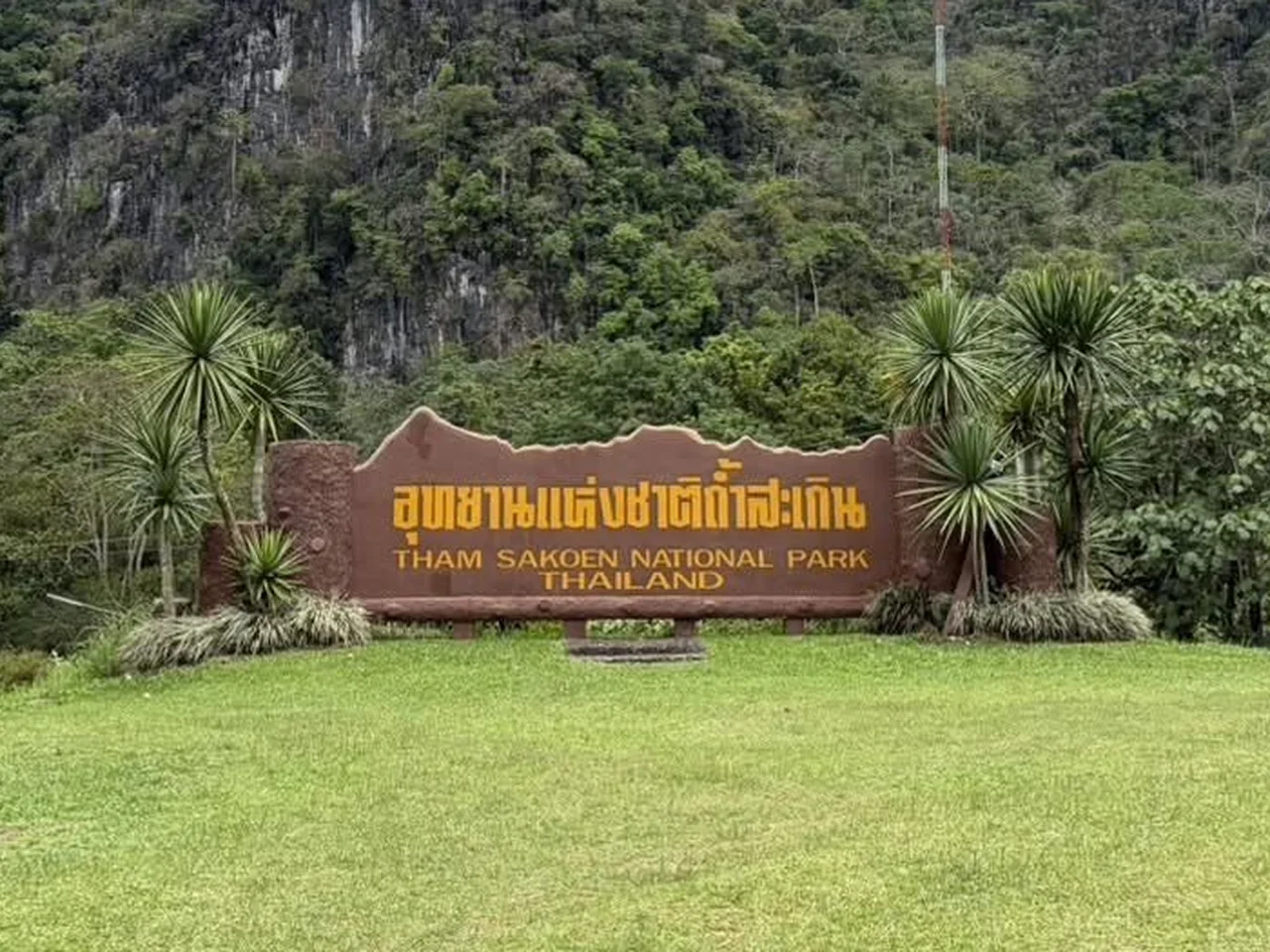 ภาพระยะใกล้ของป้ายอุทยานแห่งชาติถ้ำสะเกินบนลานหญ้า มีป่าและหน้าผาหินปูนเป็นฉากหลัง