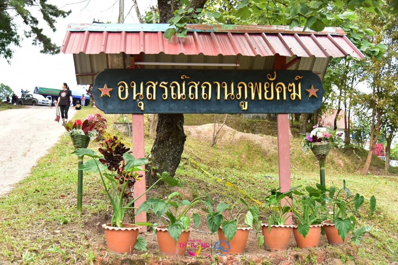 ป้ายไม้สีดำตัวอักษรสีทองเขียนว่าอนุสรณ์สถานภูพยัคฆ์ อยู่ใต้หลังคาสีแดงท่ามกลางต้นไม้