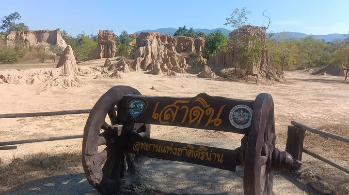ป้ายไม้เขียนว่าเสาดิน อุทยานแห่งชาติศรีน่าน โดยมีเสาดินสูงต่ำและแนวเขาเป็นฉากหลัง