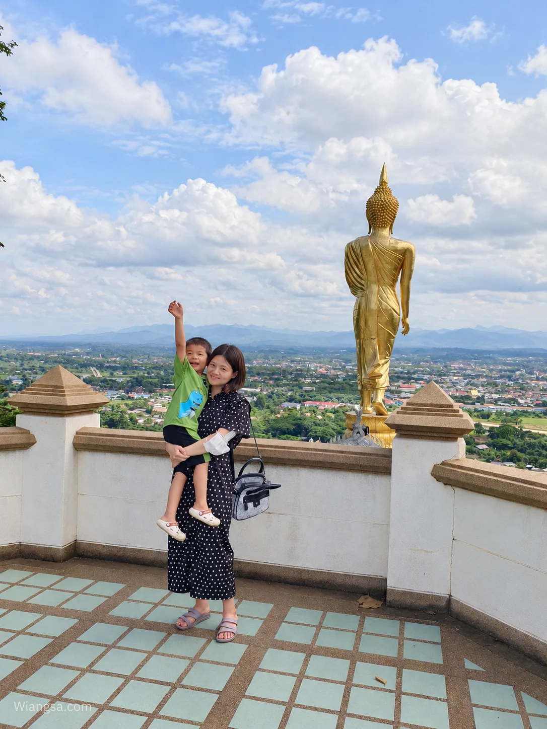 ผู้ใหญ่กำลังอุ้มเด็กที่ลานชมวิวพระธาตุเขาน้อย มีพระพุทธรูปสีทองและทิวทัศน์เมืองน่านอยู่ด้านหลัง