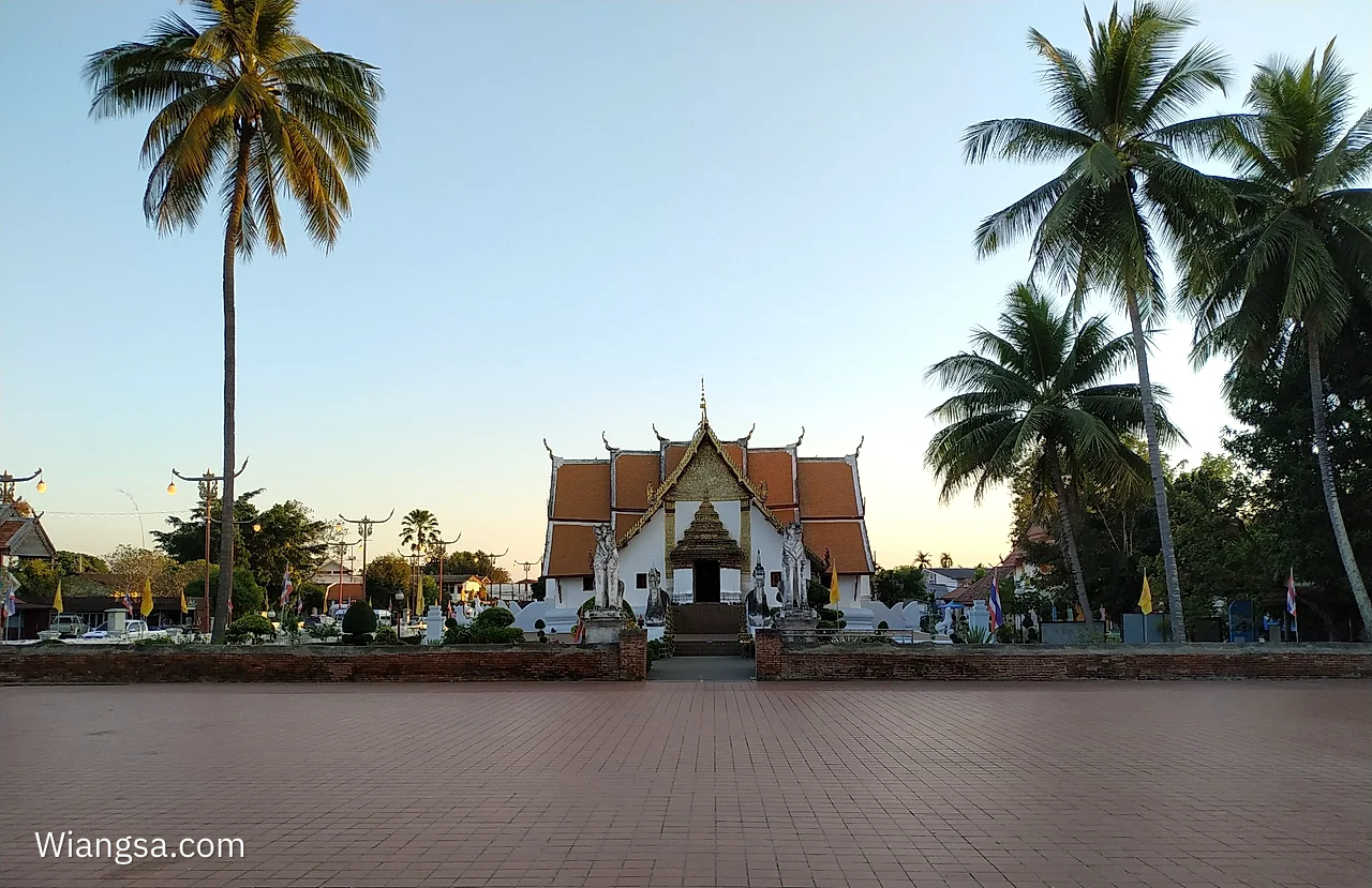 ลานกว้างด้านหน้าวัดภูมินทร์ มีแนวต้นมะพร้าวสูงล้อมอาคารวิหารหลังคาสีอิฐอยู่ตรงกลาง
